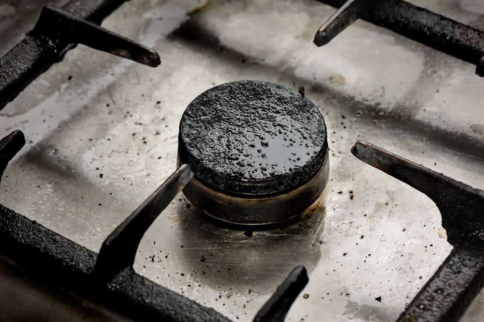 A close-up of a gas stove burner, covered in burnt and dirty residue. The metal grate around the burner also has visible signs of grime and wear, highlighting one of the common causes that can contribute to house fires. Maintain fire safety by ensuring regular cleaning and maintenance.
