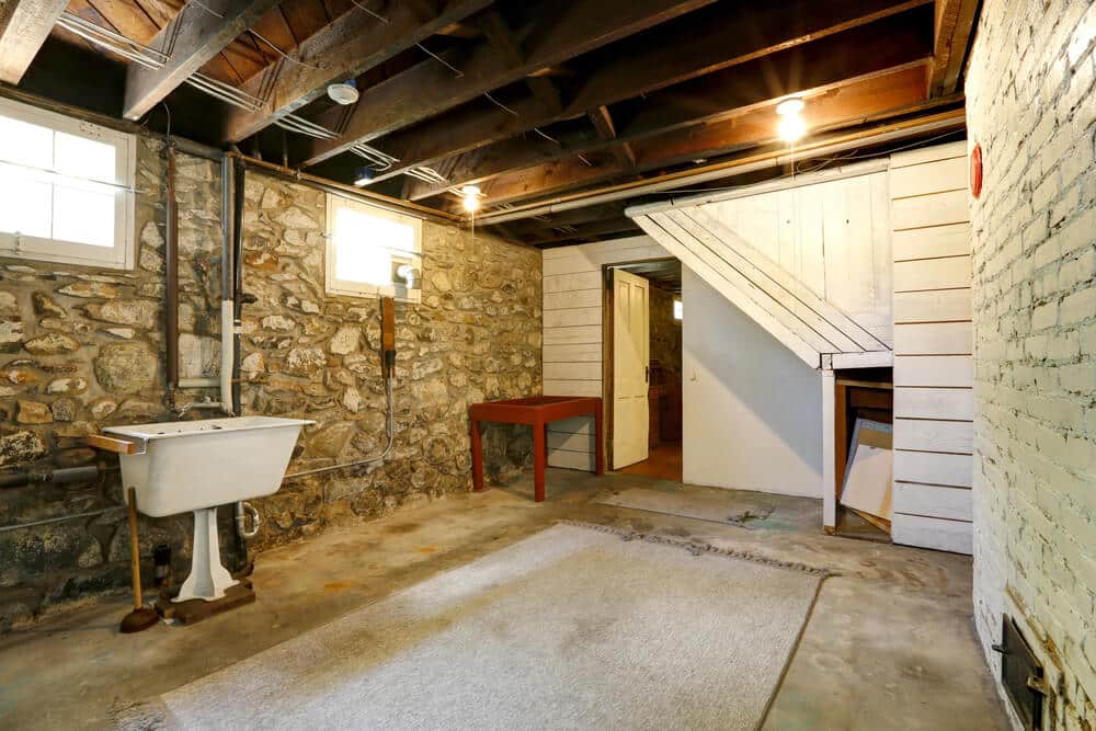 A basement with exposed wooden beams and stone walls features a sink, a small worktable, a rug, and a closet under the stairs. Alongside two small windows providing natural light, a step-by-step guide for how to remove mold from the basement walls would be handy in maintaining its charm.