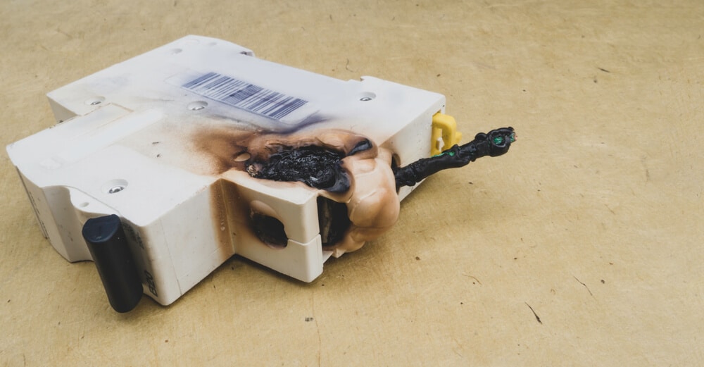 A melted and burned white electronic device with barcode, placed on a beige surface, exhibits extensive smoke damage. A cable is severely charred and damaged.
