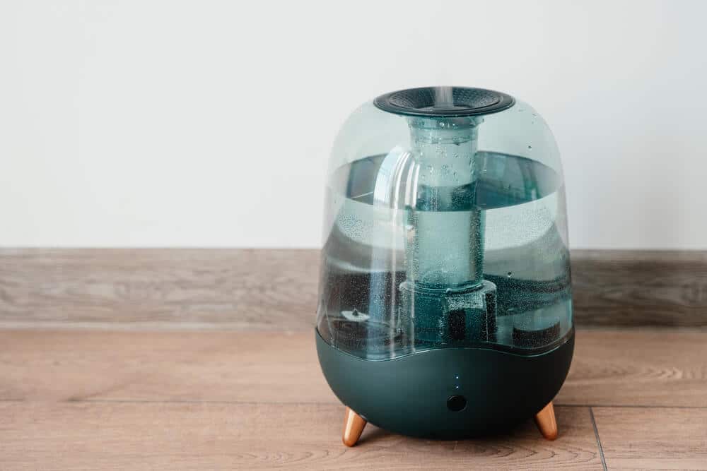 A modern, transparent humidifier with water inside stands on a wooden floor against a white wall, working efficiently to kill mold in your space.