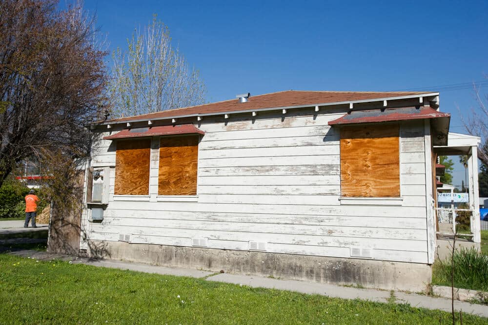 A small, weathered house with boarded-up windows stands desolate. The white paint peels sadly from its walls beneath a brown roof. For reasons of vacant house security, the windows are sealed tightly. In the background, a person in an orange shirt is barely visible.