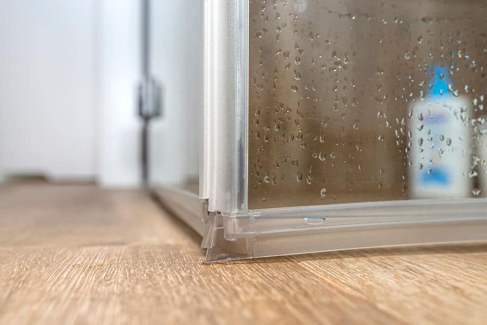 Close-up of the bottom corner of a glass shower enclosure with drops of water on the glass and a bottle of soap visible in the background. The shower sits on a wooden floor, emphasizing the importance of considering drying time to prevent potential water damage.