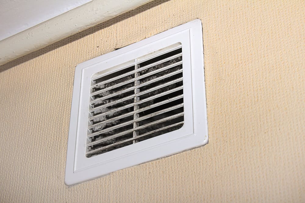 A white wall vent with dust and dirt accumulated on its slats against a beige wall, highlighting the need for proper crawl space ventilation.