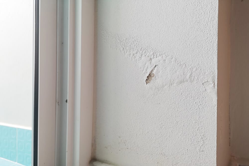 A close-up of a white wall with a crack and a small hole near a window frame. The wall appears textured, with slight discoloration suggesting damp marks on walls around the damaged area.