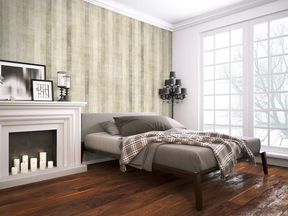 A modern bedroom features a grey bed with a plaid blanket, a fireplace with candles, framed wall art, and a large window. If you need to remove mold from the plywood subfloor, it's important to address it promptly for a clean and healthy living space.