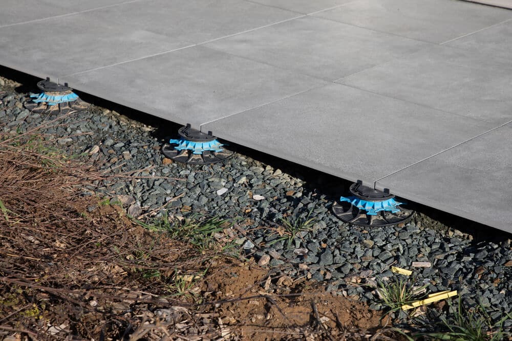 Close-up of outdoor tiles being installed on a raised pedestal system over a gravel and dirt base. The blue pedestal supports are visible beneath the tiles, ensuring proper patio slope for effective house drainage.