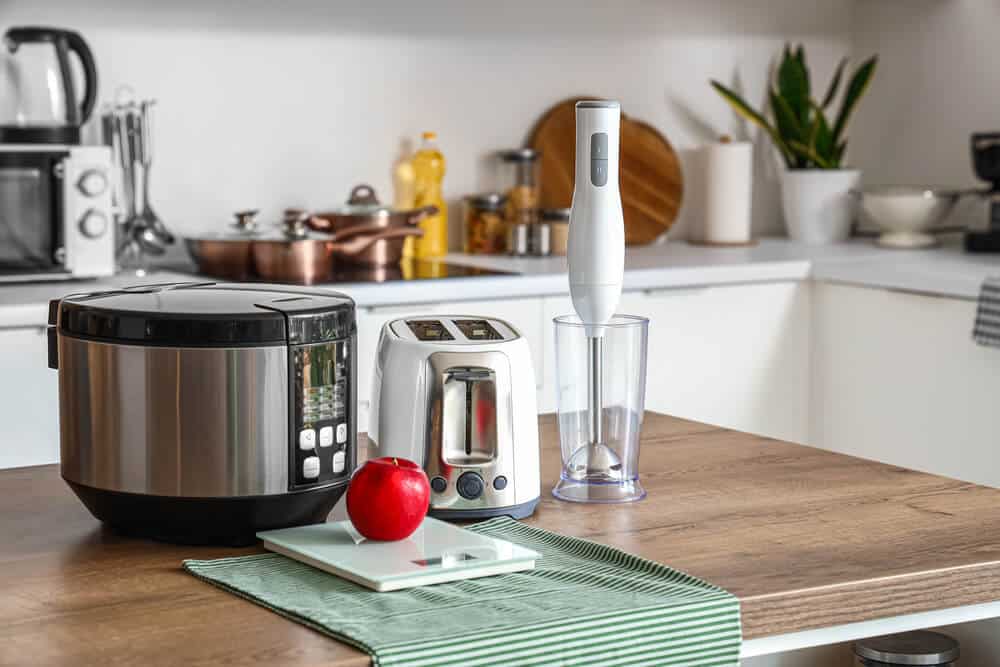 A modern kitchen counter with a slow cooker, toaster, immersion blender with cup, cutting board with red apple, and a green-striped cloth. Remember to follow fire safety guidelines when using any electrical appliance to prevent hazards.