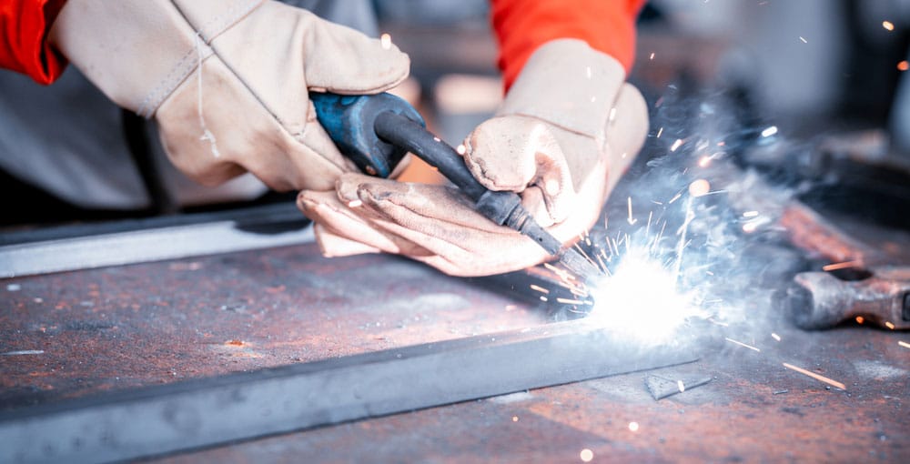 Close-up of a person welding metal with bright sparks flying, highlighting the importance of safety. They are wearing protective gloves and an orange sleeve, emphasizing precautions against fire risks in the factory.