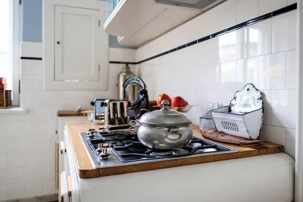 A kitchen counter with a stove, a stainless steel pot with a lid, a toaster, a grater, and a bowl of apples. White tiled walls and cabinets are in the background. Ensure to avoid common hazards by organizing sharp tools safely.