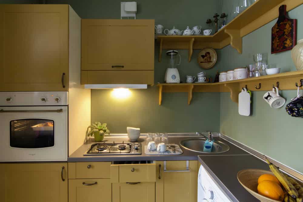 A small kitchen with yellow cabinets, a gas stove, oven, sink, blender, and shelves with teapots, glasses, and dishes. A bowl of oranges is on the counter. To ensure workplace safety and prevent fire hazards, the area is equipped with a fire extinguisher within easy reach.