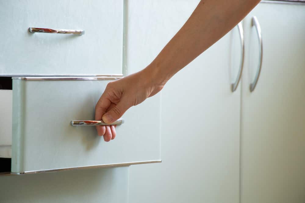 A person opens a light-colored cabinet drawer, revealing the handle and other nearby door handles. The scene hints at the need for kitchen cabinet repair after water damage.