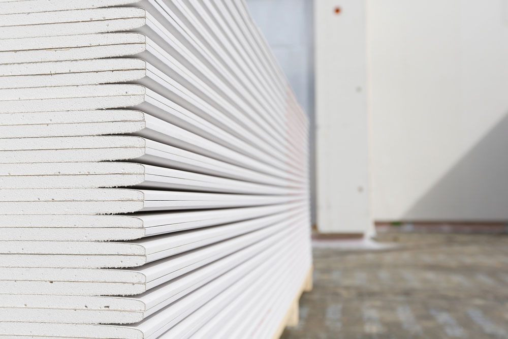 Stack of white gypsum board sheets neatly piled outdoors on a paved surface, with a building wall in the background.