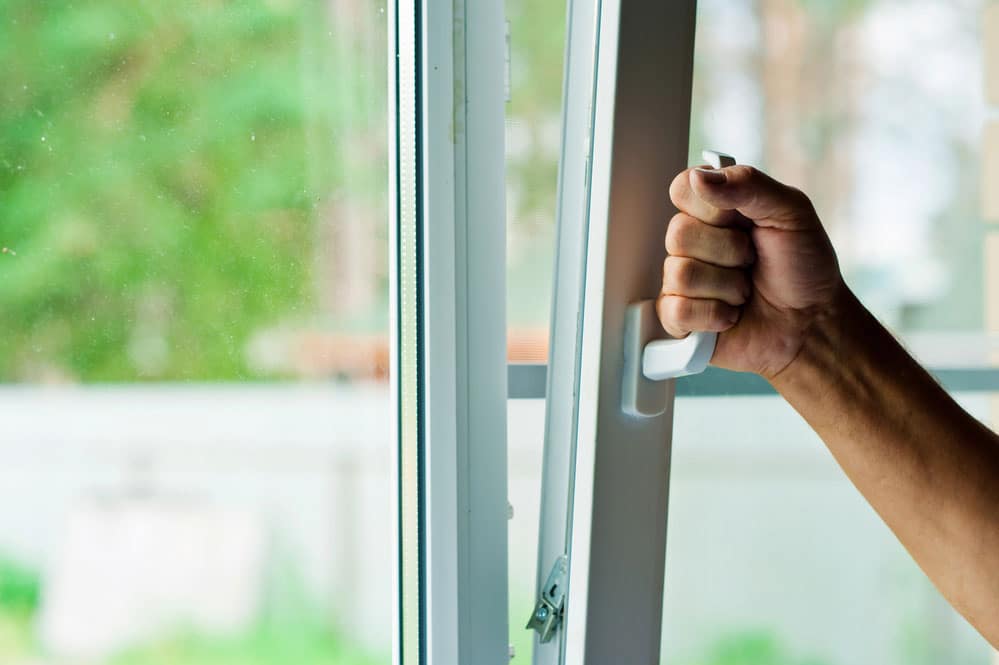 A hand is gripping the handle of a white window, which is partially open, revealing a blurred outdoor background—one of the effective ways to ensure storm safety and protect windows.