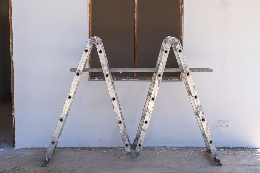 A double-sided ladder is positioned in an "M" shape against a wall with an open window frame and a power outlet, ensuring the area matches the acceptable moisture percentage for drywall.