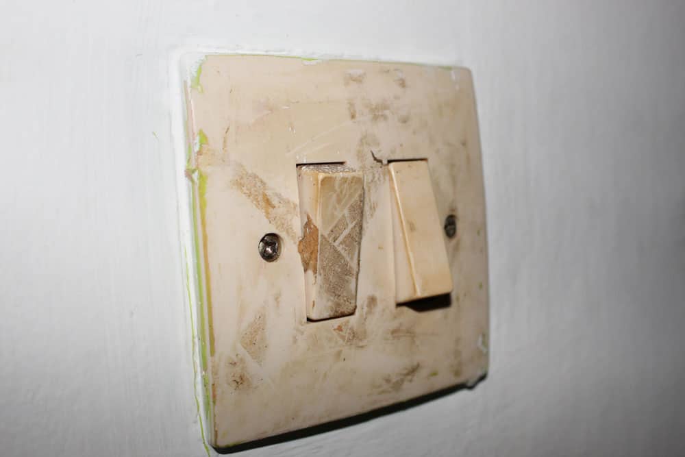 A dirty and worn light switch plate on a white wall with two toggle switches. The discolored plate, with chipped paint around the edges, can be a potential cause of electrical fires if not addressed promptly. Fire safety should be a priority in maintaining such fixtures.