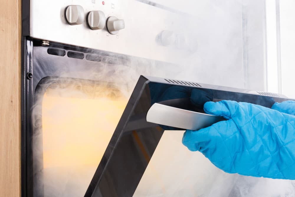 Person wearing blue oven mitts opening an oven door with smoke coming out, looking concerned as if wary of inhaling the thick smoke, which could make them sick.